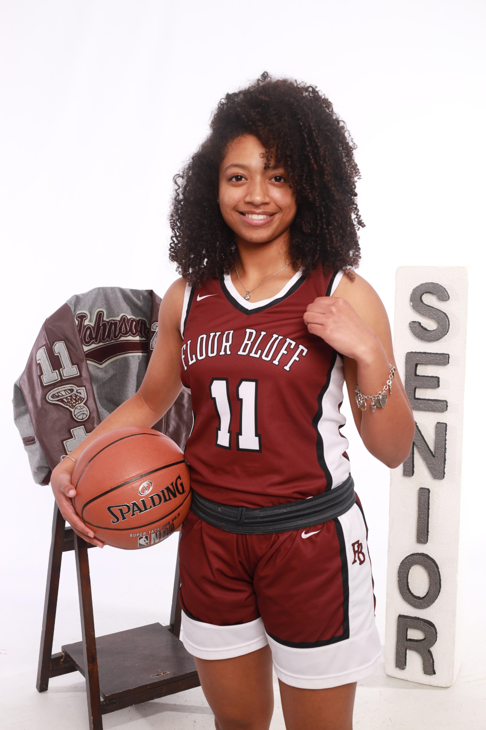 Teenage girl posing with a basketball in her uniform. In the background is her letterman's jacket and stacked letter blocks reading "senior".