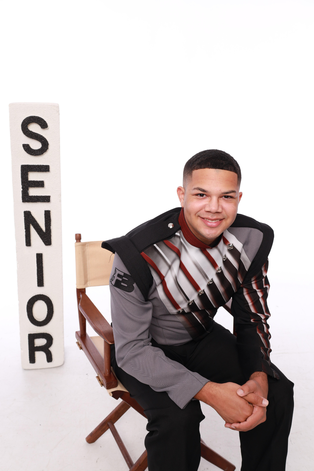 Teenage boy in a marching band uniform with a stacked letter block sign reading "senior".