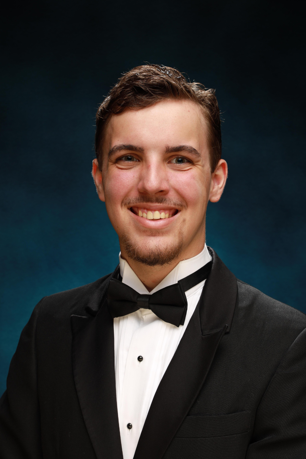 Teenage boy wearing a black tie and tux against a blue background.