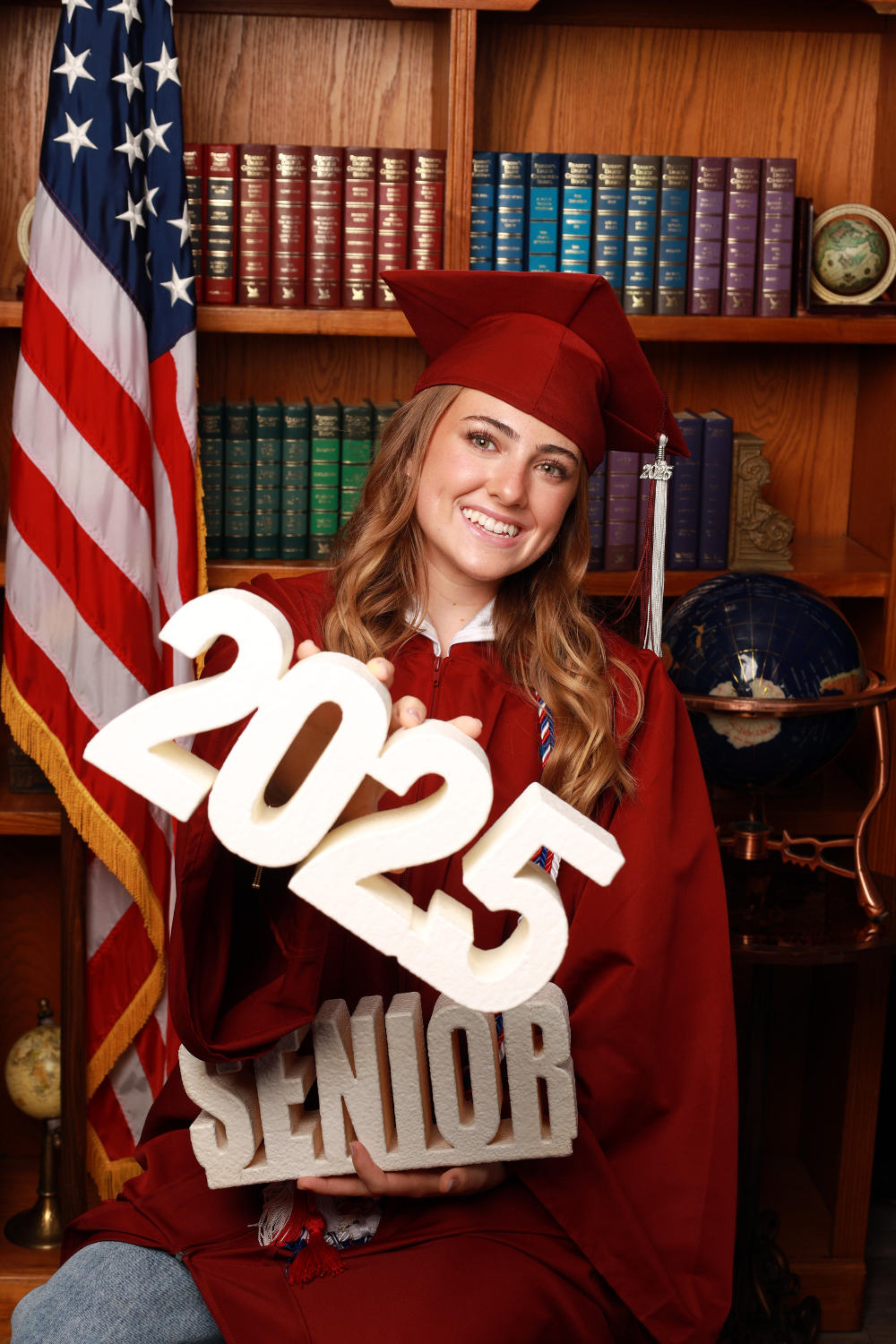 Female graduate wearing a red graduation cap and gown holding a "2025 senior" sign.