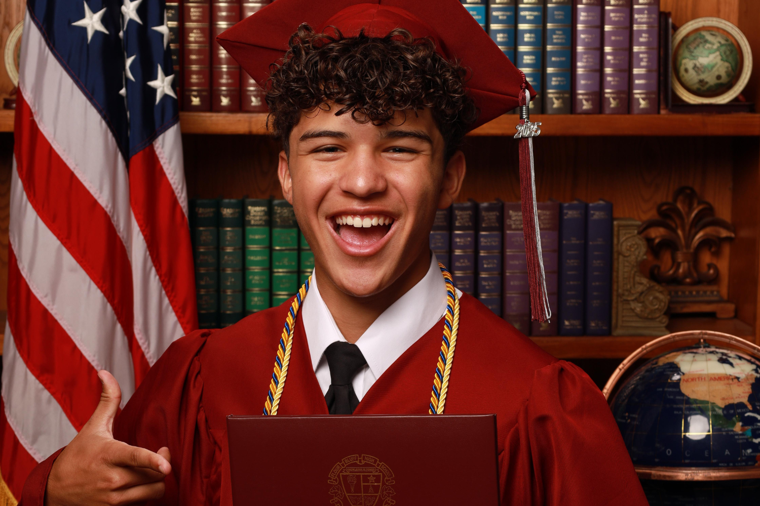 Teenage boy wearing a maroon graduation cap and gown, excitedly pointing to a diploma.