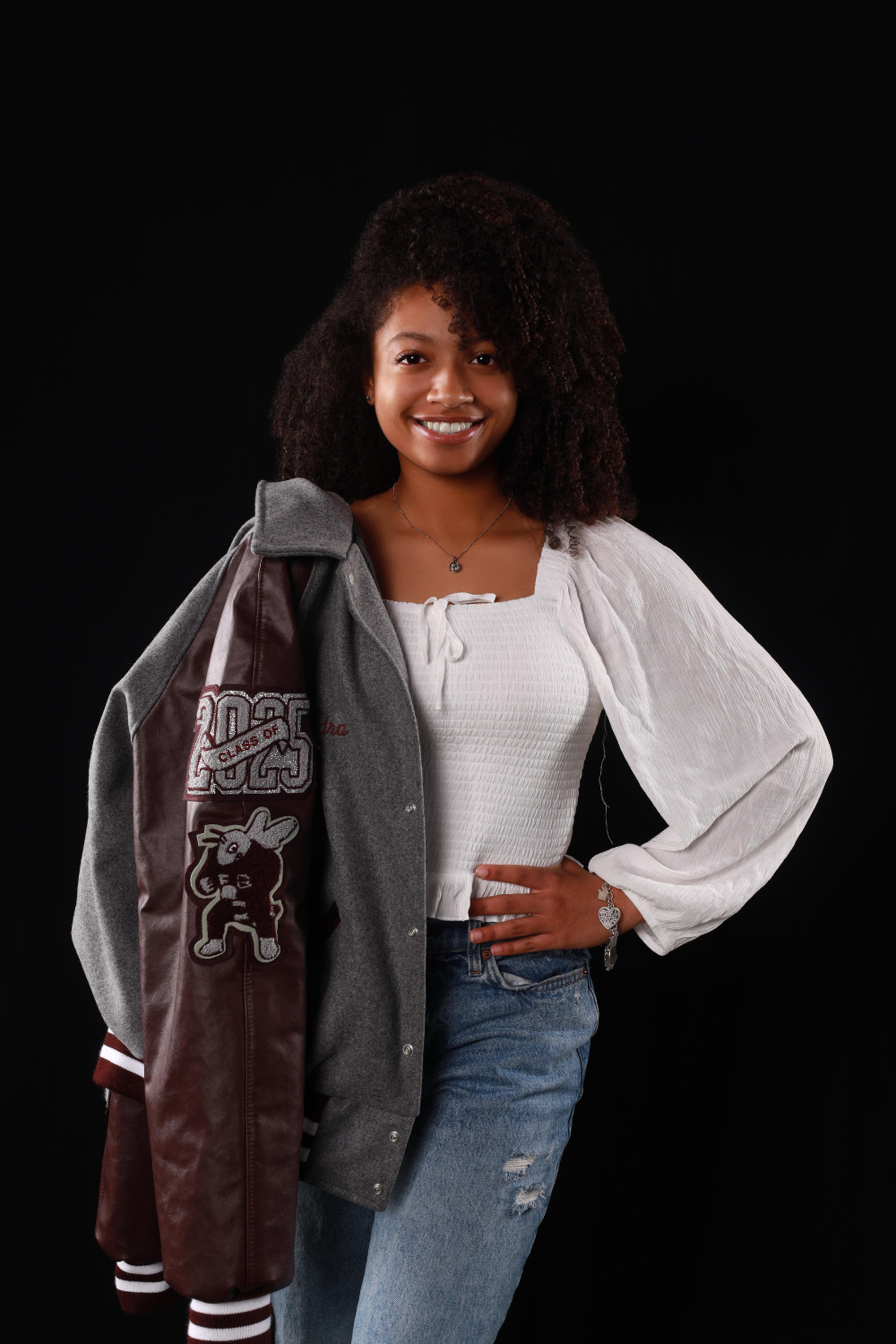 Senior portrait of a teenage girl wearing a white top and blue jeans holding her letterman's jacket over one sholder.