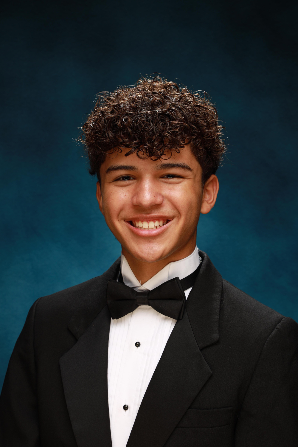 Formal portrait of a teenage boy wearing a black tux and bow tie against a blue backdrop.