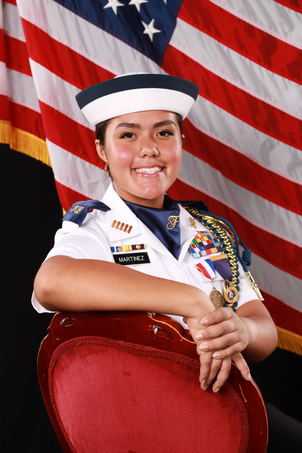 Senior portrait of a teenage girl sitting in a red chair, wearing a military uniform, with the American flag in the background.