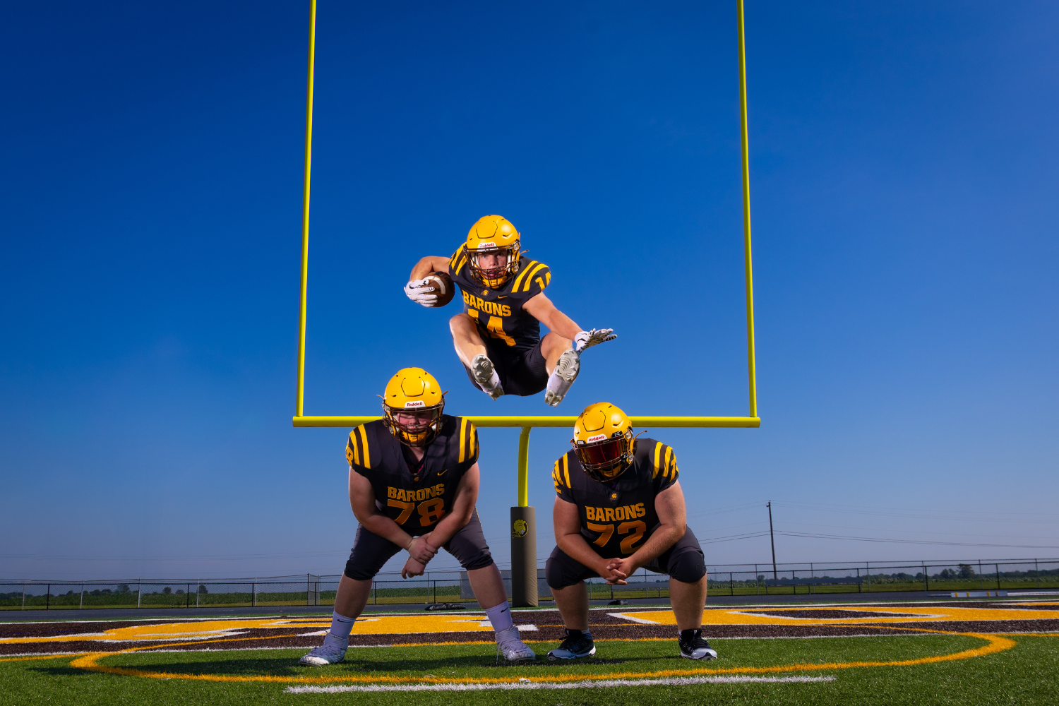 Two highschool football players leaned forward while a third jumps over them.